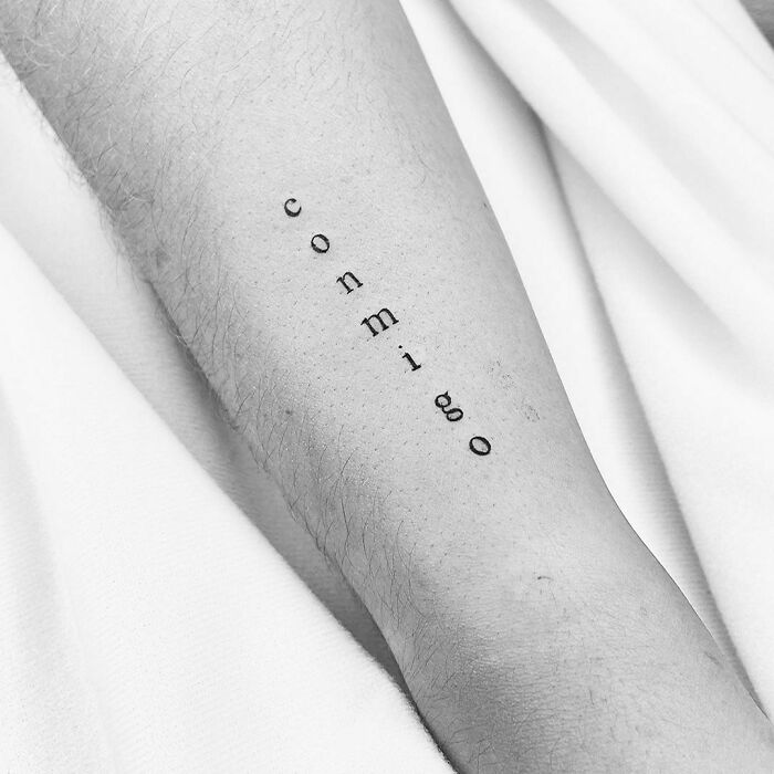 Vertical word tattoo on forearm spelling "conmigo," showcasing a simple design idea for new ink enthusiasts.