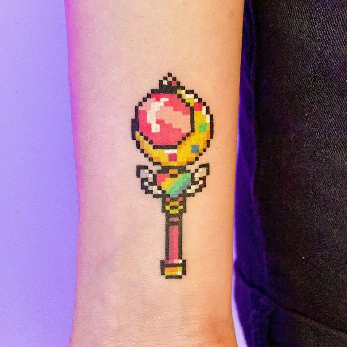 Pixelated cartoon tattoo of a colorful magical staff on forearm.