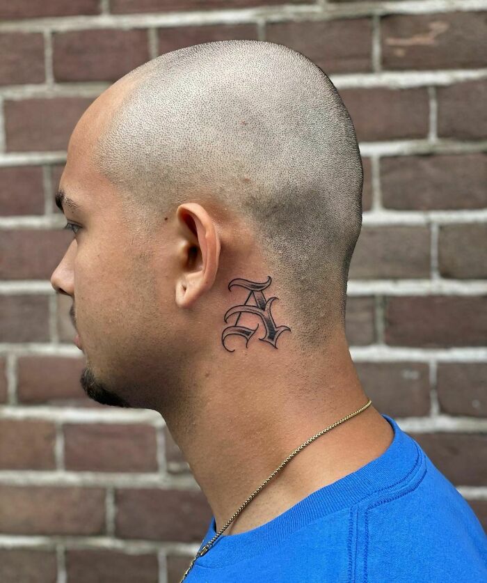 "Man with shaved head displaying a bold letter tattoo behind ear against a brick wall."
