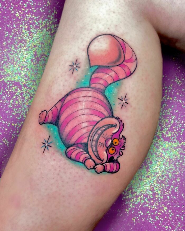 Colorful cartoon tattoo of a pink-striped cat Cheshire with a big grin on a person's arm, surrounded by sparkles.