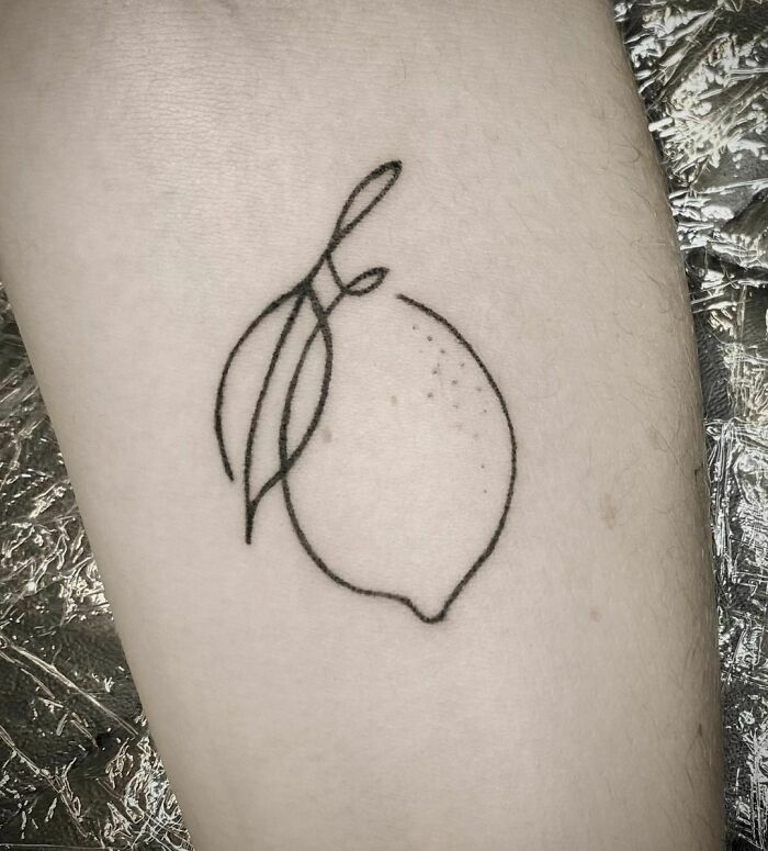 Single-line tattoo of a stylized lemon with leaves on skin.