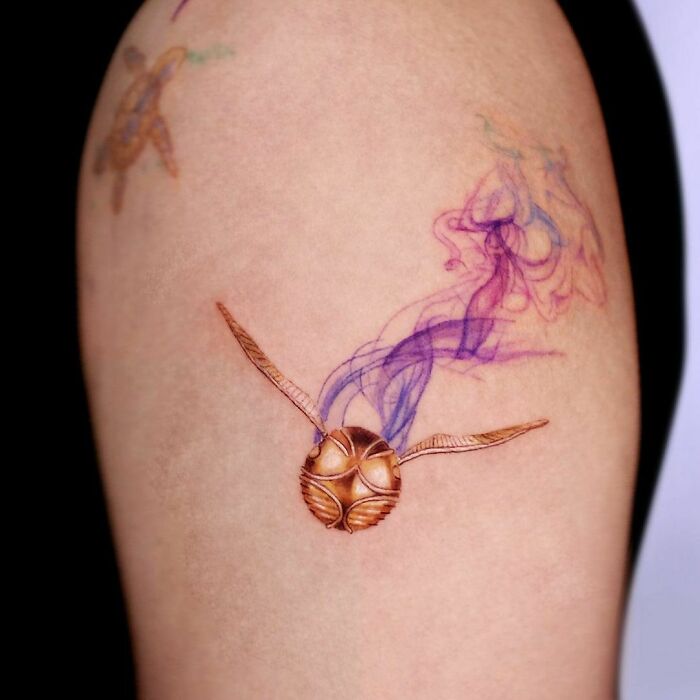 Golden Snitch tattoo on arm with colorful smoke design, ideal for Harry Potter fans.