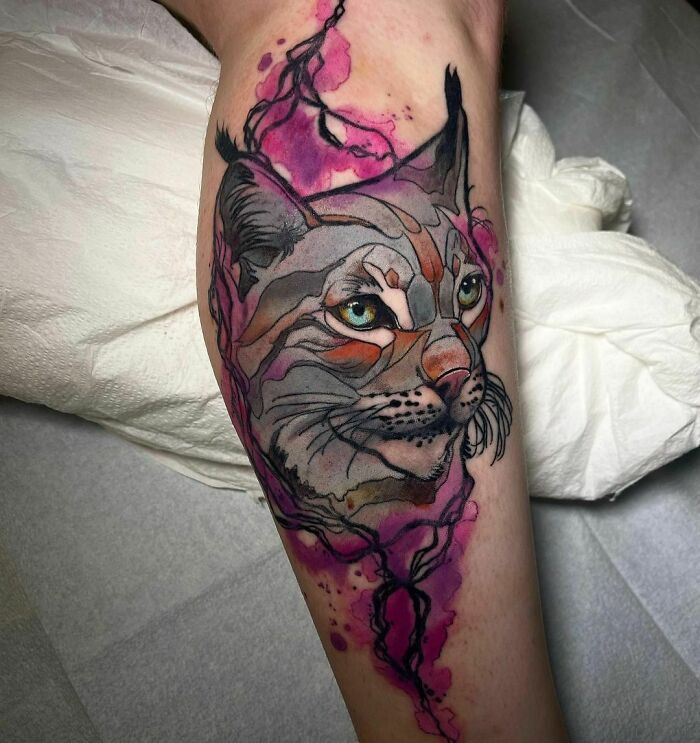 Watercolor tattoo of a lynx with vibrant pink and purple splashes on an arm.