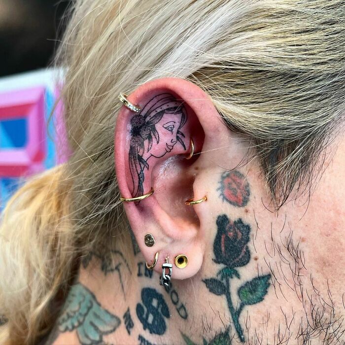 Ear tattoo featuring a detailed feminine face, surrounded by multiple gold earrings and additional neck tattoos.