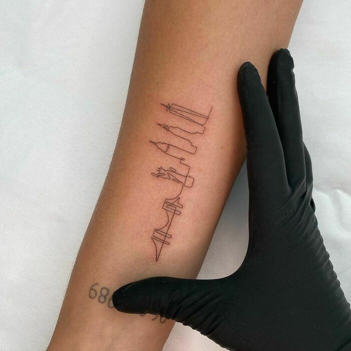 Fine-line single-line tattoo of a city skyline on a forearm, professionally done with a glove-covered hand beside it.