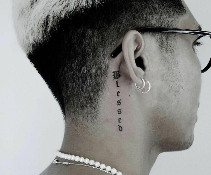 Close-up of a man's ear tattoo reading "Blessed," with earrings and short hair, highlighting ear tattoo design.