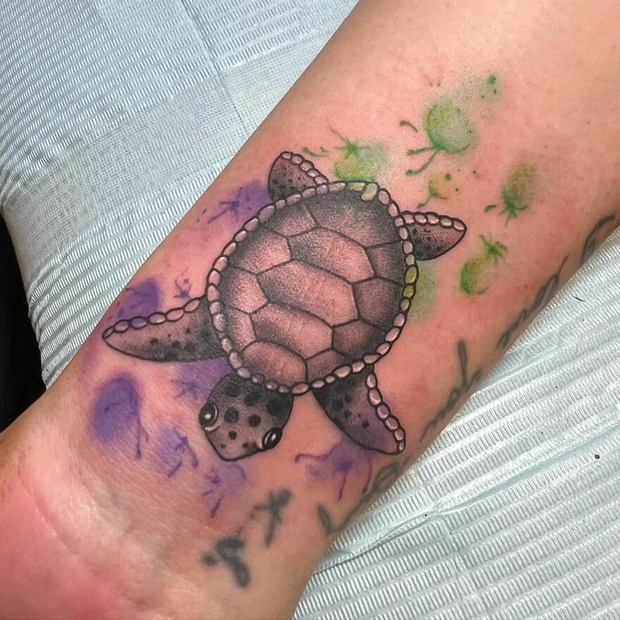 Watercolor turtle tattoo design on forearm with vibrant green and purple splashes, showcasing artistic beauty.