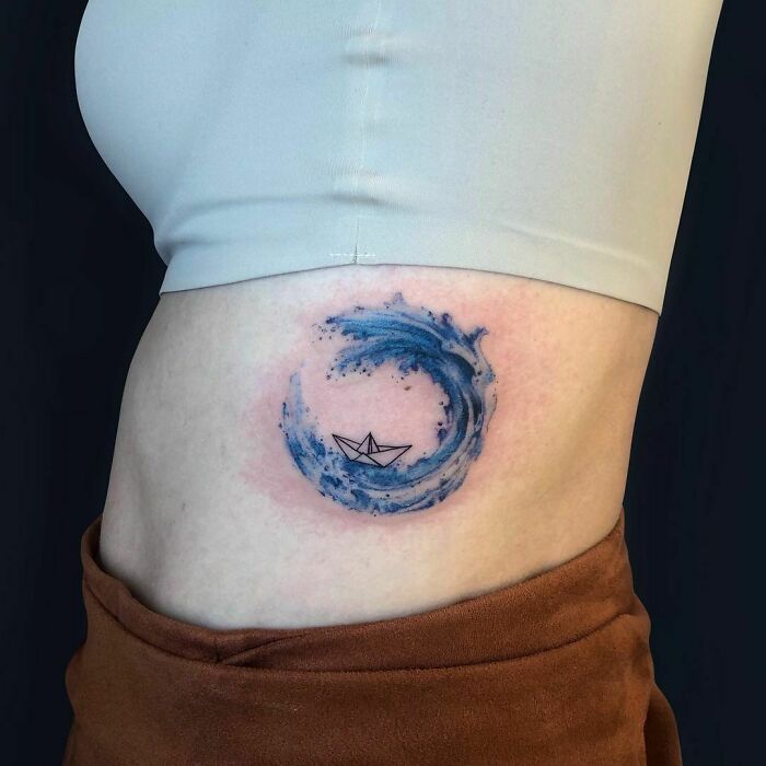 Watercolor tattoo of a blue wave with a paper boat on a person's side, showcasing ethereal artistry.