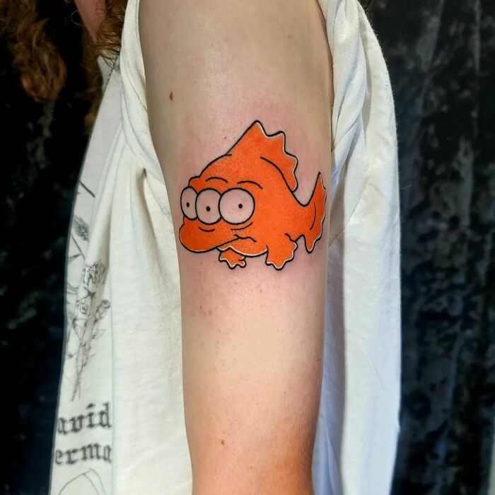 Cartoon tattoo of a three-eyed orange fish on a person's arm, inspired by animated shows.