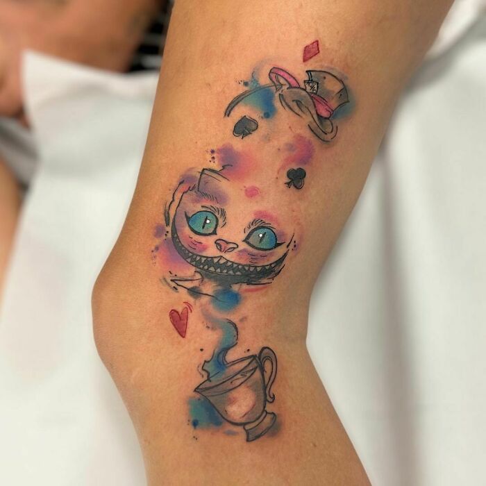 Watercolor tattoo featuring a whimsical Cheshire cat design with vibrant blues and pinks, creating an ethereal look.