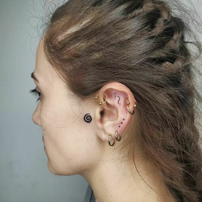Close-up of ear with multiple tattoos and piercings, featuring a spiral design and Saturn symbol.