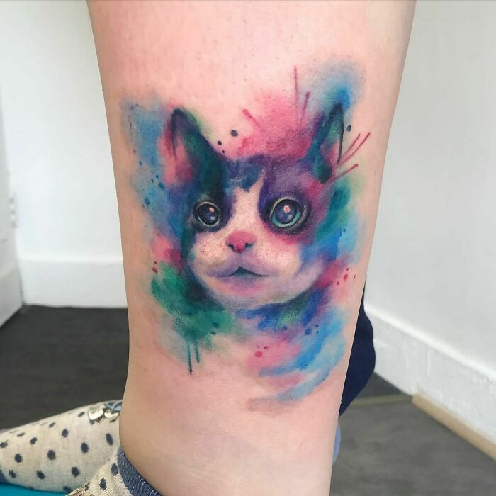 Watercolor tattoo of a cat's face with colorful, ethereal splashes on a person's leg.