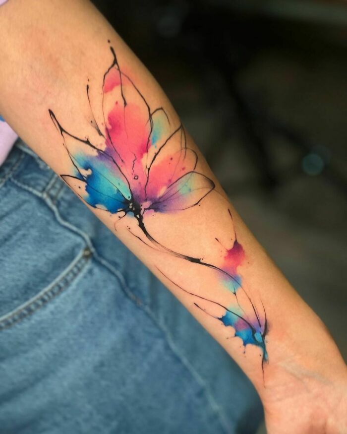 Colorful watercolor tattoo of a butterfly design on a forearm, blending pink, blue, and purple tones.