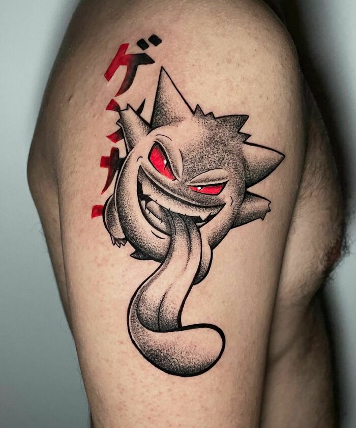 Cartoon tattoo of a grinning character Gengar with red eyes and Japanese characters on someone's arm.