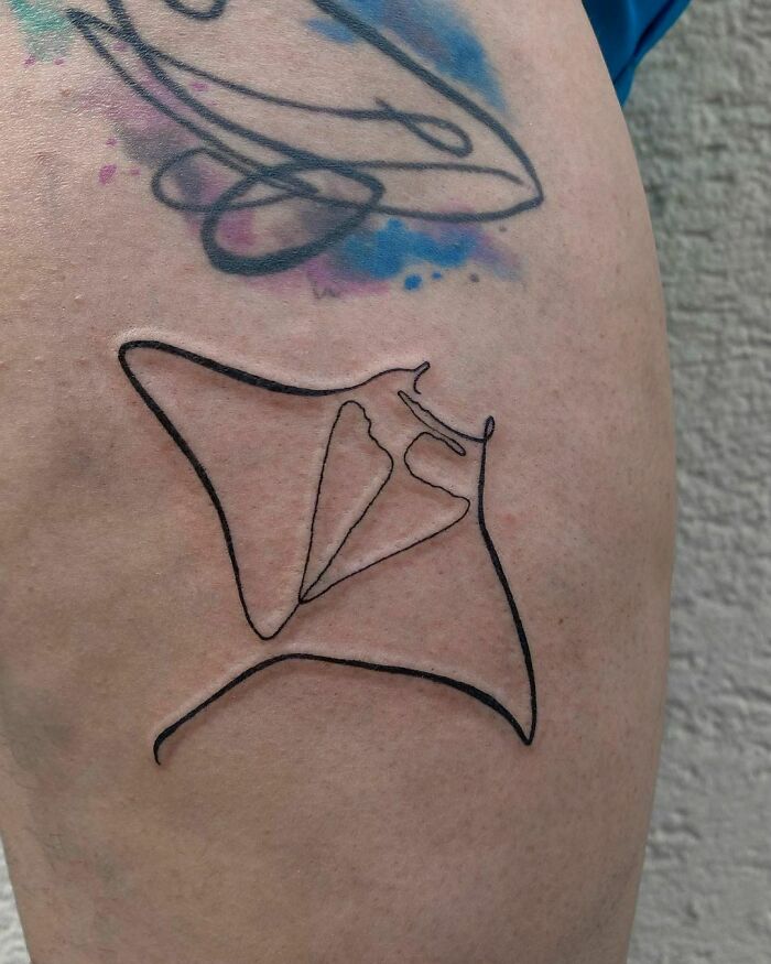 Single-line tattoo of a manta ray on skin with colorful ink accents in the background.