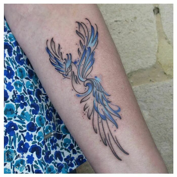 Blue and black phoenix watercolor tattoo on forearm, showcasing ethereal design.