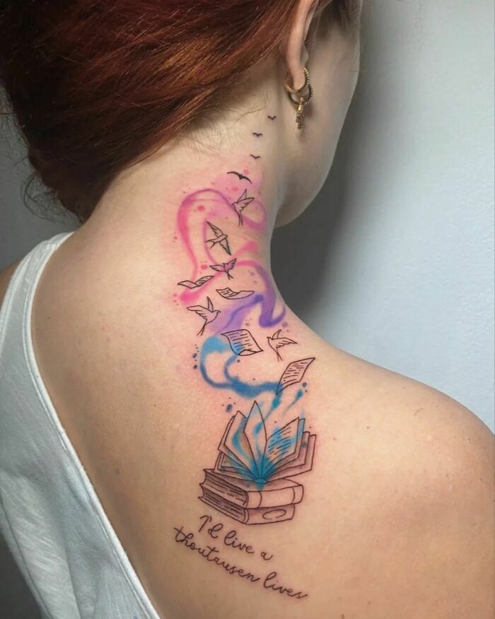 Watercolor tattoo of books and flying pages on a woman's back, blending vibrant pink and blue inks.