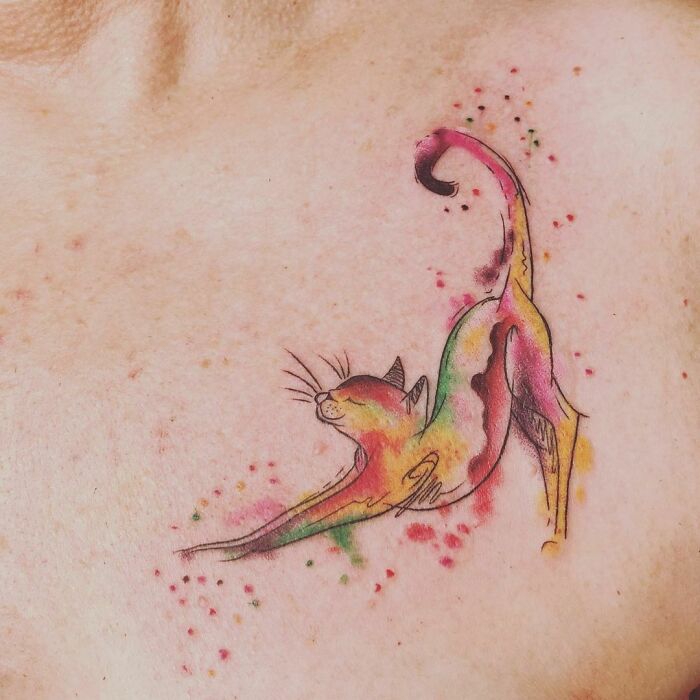 Watercolor tattoo of a stretching cat in vibrant, ethereal hues on skin.
