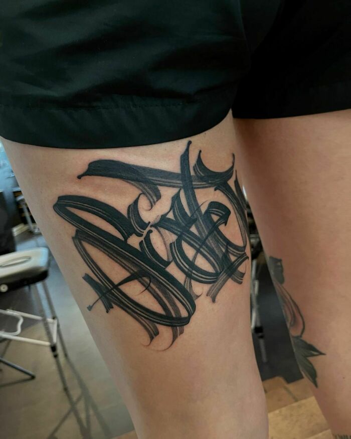 Tattoo featuring intricate black lettering on a person's thigh, showcasing unique word design.
