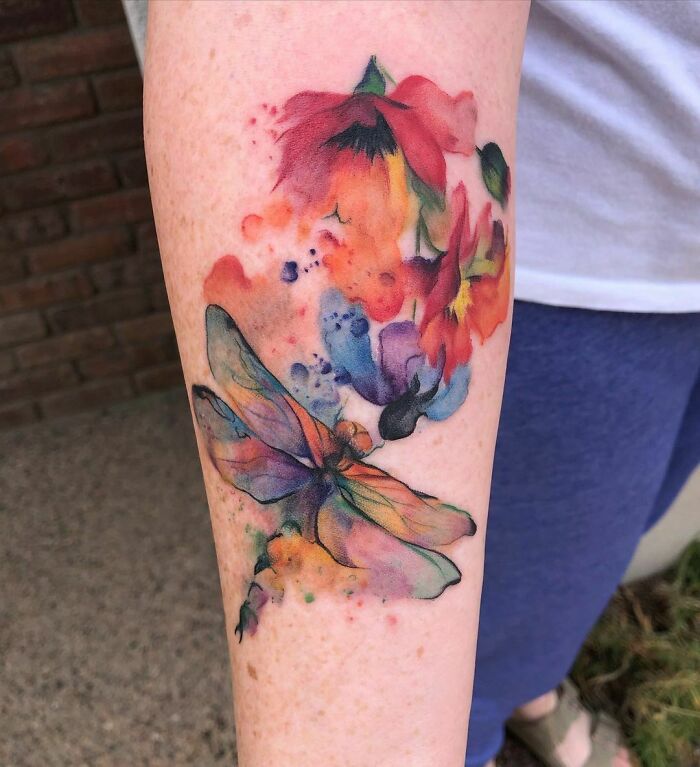 Watercolor tattoo featuring a colorful dragonfly and flowers on forearm.