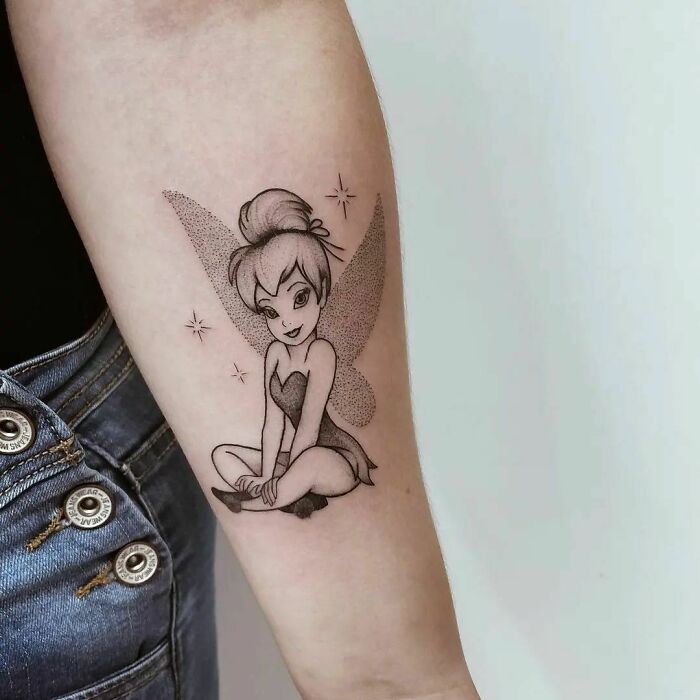 Cartoon tattoo of Tinker Bell with crossed legs on forearm, evoking nostalgia.