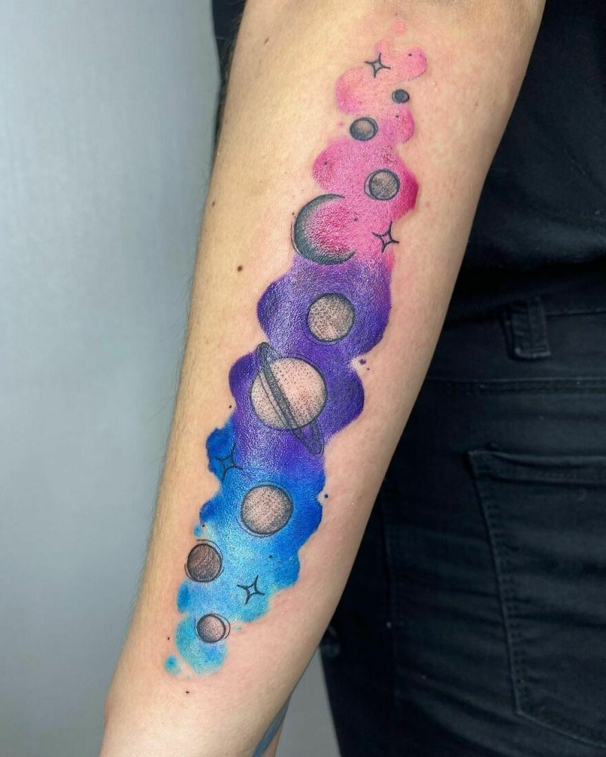 Watercolor tattoo of colorful planets and stars on an arm, blending pink, purple, and blue hues beautifully.
