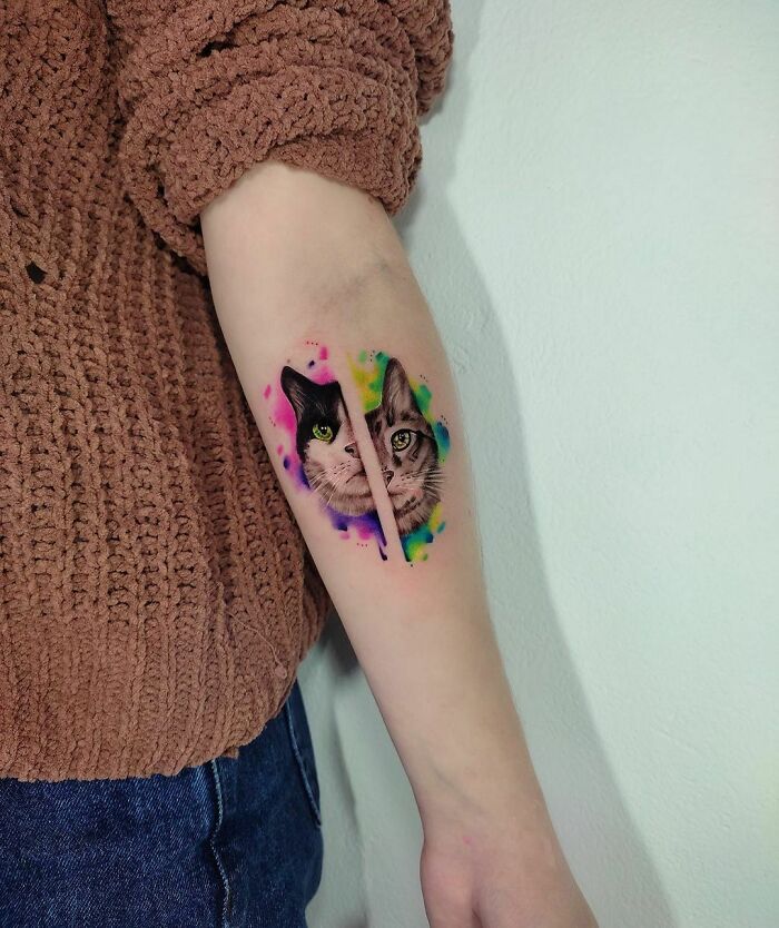 Watercolor tattoo of two cat faces on an arm, surrounded by colorful splashes, showcasing beautiful inkspiration.