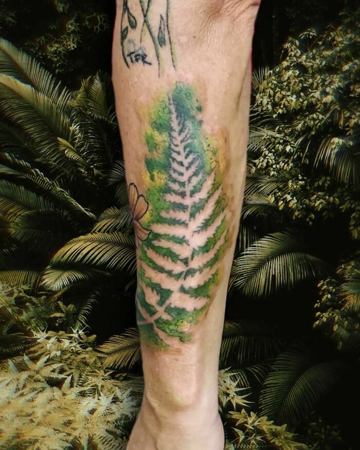 Watercolor tattoo of a green fern on a leg amidst a lush background, showcasing beautiful inkspiration.