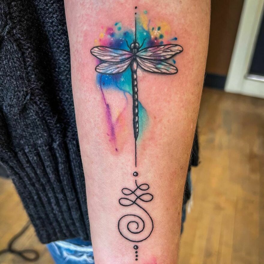 Watercolor tattoo of a dragonfly with colorful splashes on an arm.