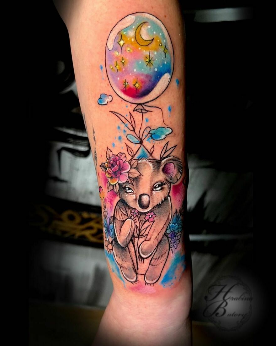 Watercolor tattoo of a koala with flowers and a moon balloon in vibrant colors on an arm.