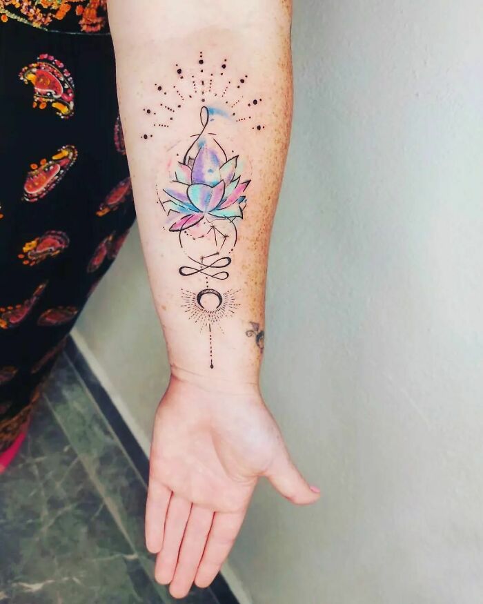 Watercolor tattoo on forearm featuring a colorful lotus with geometric and celestial designs.