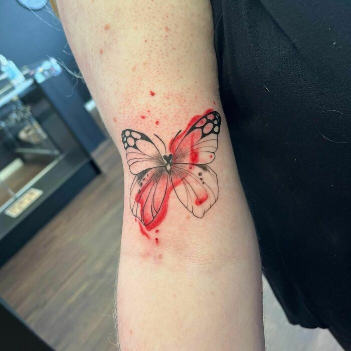 Watercolor tattoo of a butterfly with red accents on an arm. Ethereal and artistic design.