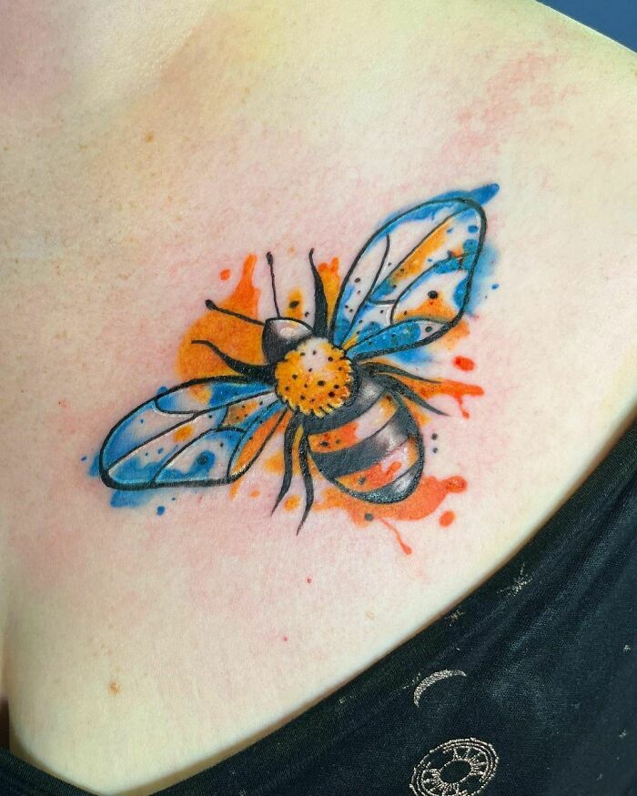 Colorful watercolor bee tattoo on skin with vibrant blue and orange splashes.