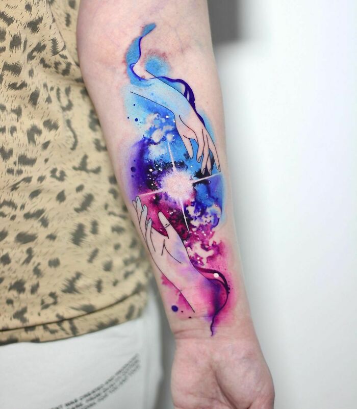 Ethereal watercolor tattoo of abstract hands with vibrant cosmic colors on arm.