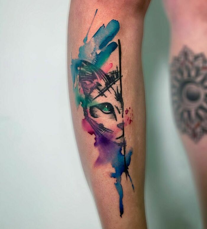 Watercolor tattoo of a cat design on a person's leg, featuring vibrant splashes of blue, green, and purple ink.