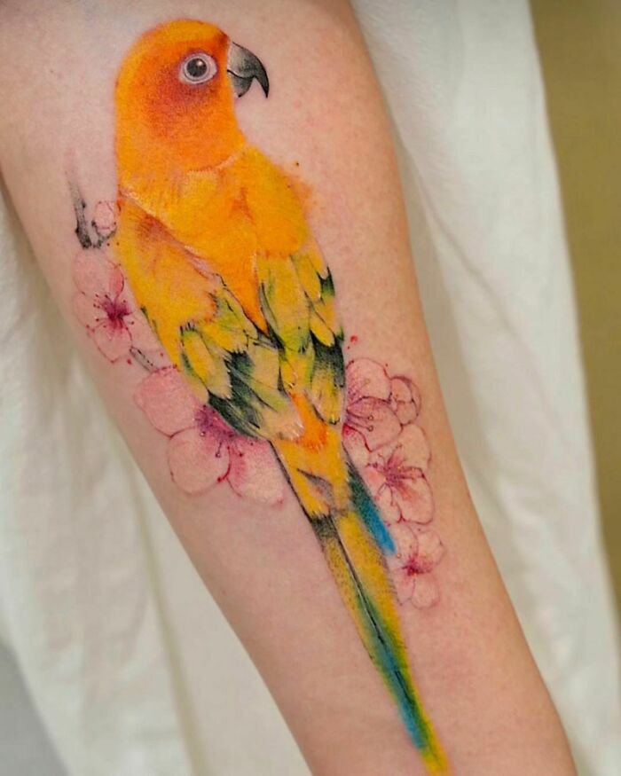 Watercolor tattoo of a vibrant parrot with pink flowers on an arm, showcasing ethereal art.