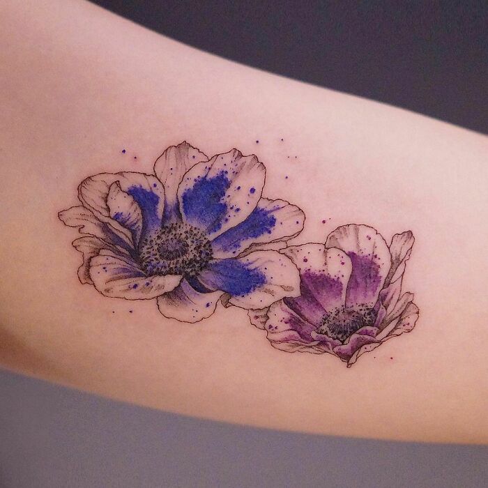 Two ethereal watercolor flower tattoos in purple and blue on a forearm.