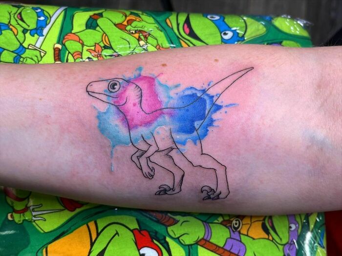 Watercolor tattoo of a dinosaur in blue and pink hues on a forearm, set against a colorful background.