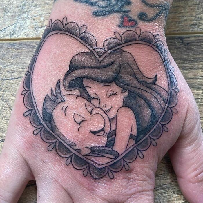 Cartoon tattoo of two characters embracing, surrounded by a heart and lace design on a hand.
