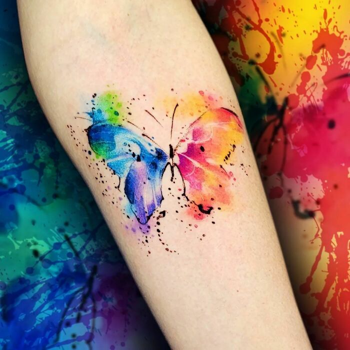 Colorful watercolor butterfly tattoo on forearm, blending vibrant blue, pink, and yellow hues in ethereal style.