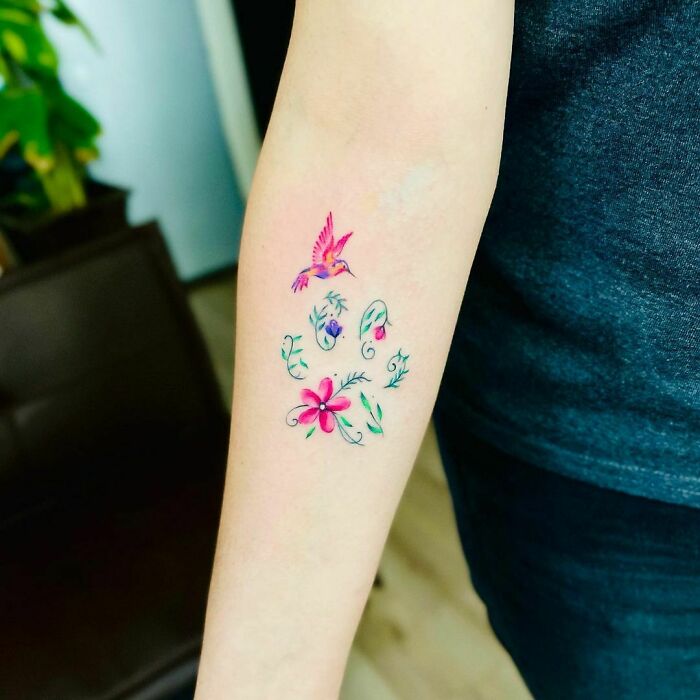 Colorful watercolor tattoo of a bird and floral design on a person's forearm.