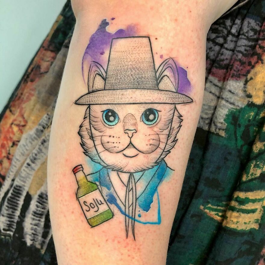 Watercolor tattoo of a cat in a hat holding a soju bottle, with vibrant purple and blue splashes on an arm.