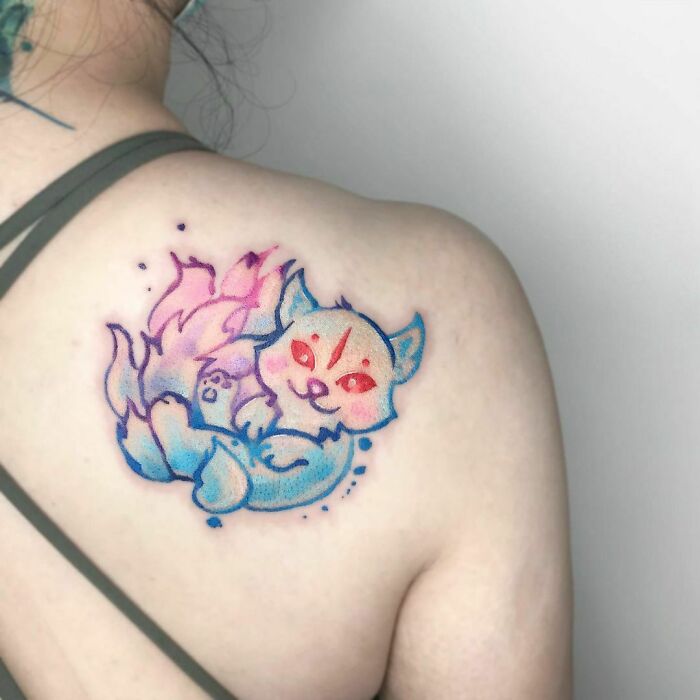 Colorful watercolor tattoo of a playful fox on a person's shoulder.