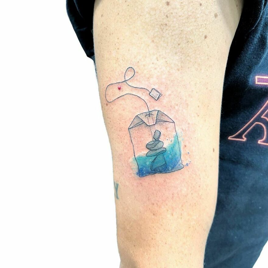 Watercolor tattoo of a tea bag with water and stones, inked on a person's arm.