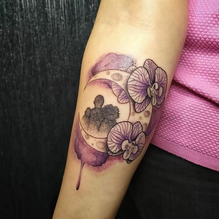 Watercolor tattoo of a crescent moon with purple flowers and silhouettes on an arm.