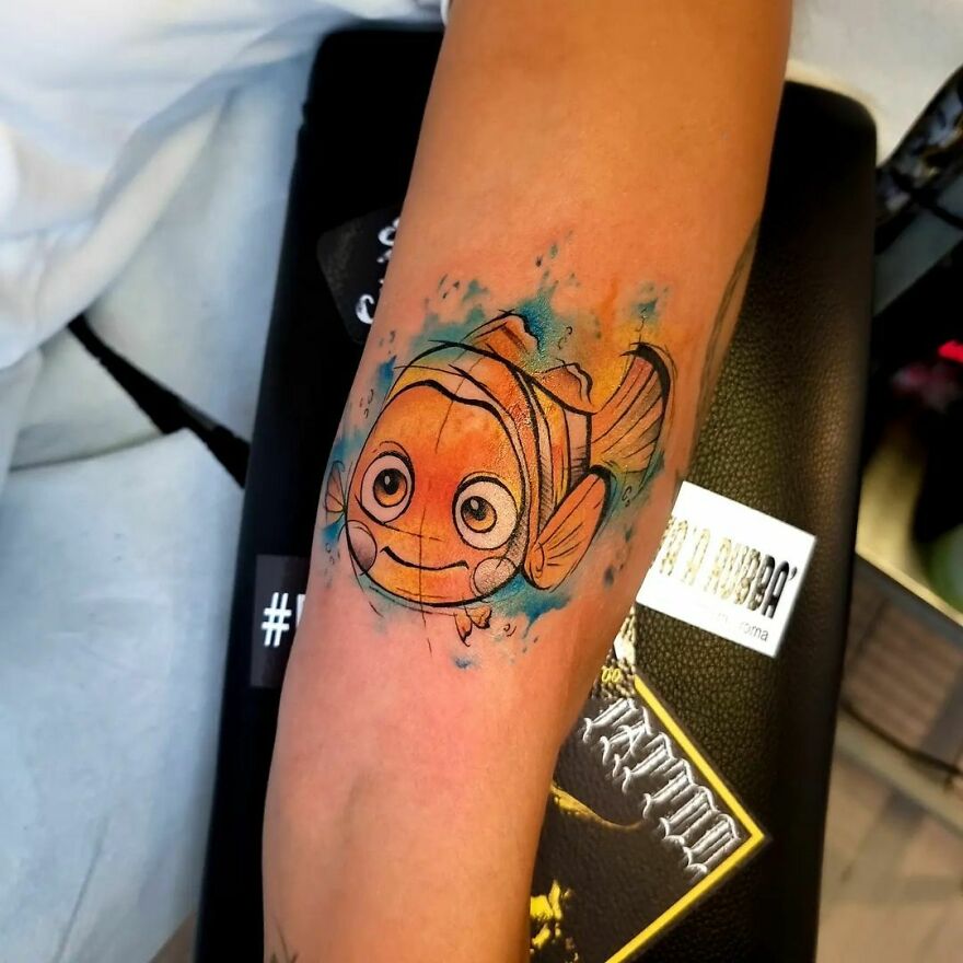 Watercolor tattoo of a cartoon fish on forearm, featuring vibrant orange and blue hues.