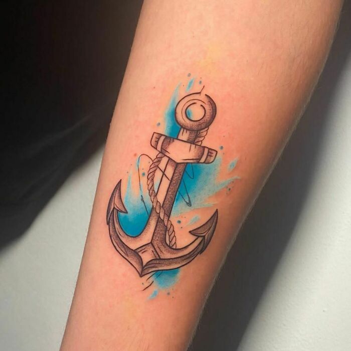 Watercolor tattoo of an anchor with blue splashes on a forearm.