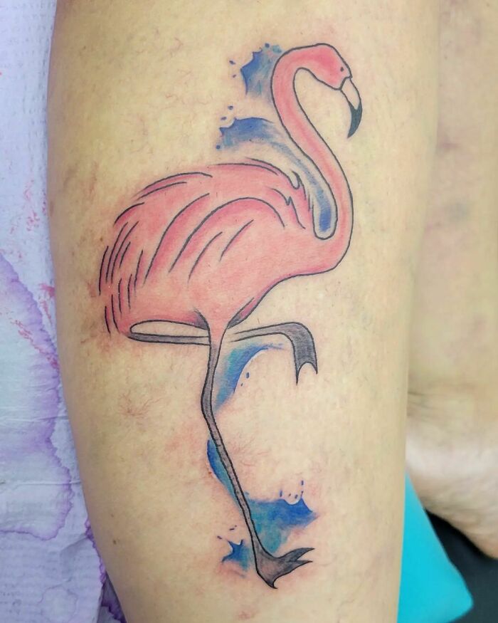 Watercolor tattoo of a flamingo with splashes of blue ink on skin.