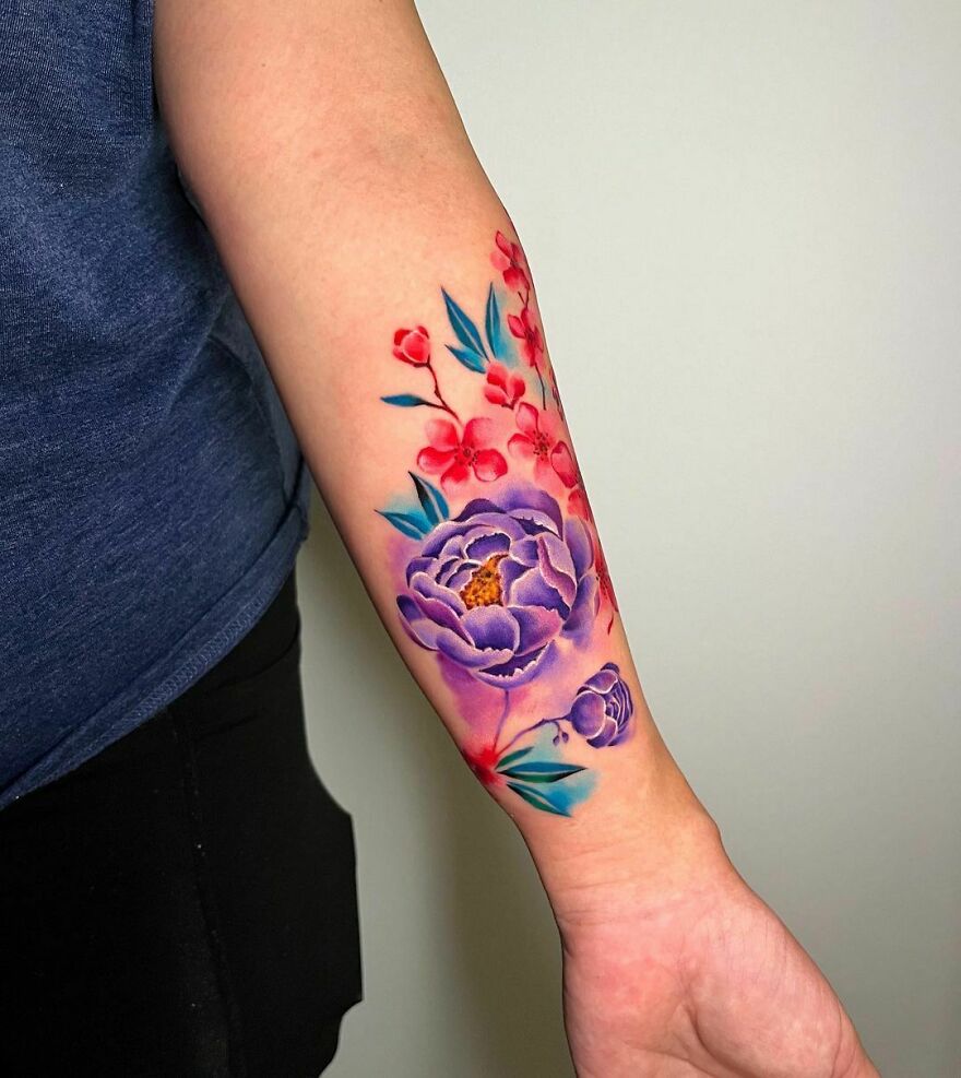 Colorful watercolor tattoo featuring purple flowers and red blossoms on a person's forearm.