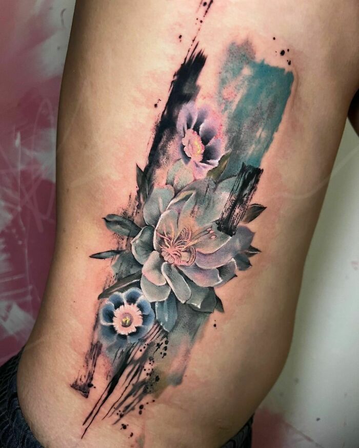 Watercolor tattoo of ethereal flowers in blue and pink tones on skin, showcasing artistic brushstroke effects.